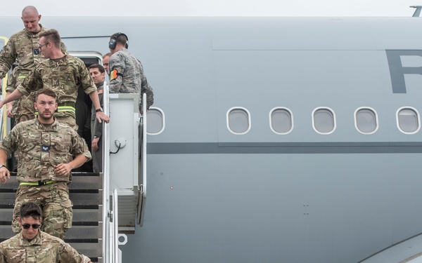 Military personnel in camouflage uniforms are disembarking from a large grey military aircraft via a mobile staircase. The aircraft displays partial lettering 'RO' on its fuselage, consistent with a Royal Air Force transport aircraft. Approximately six to seven individuals are visible descending or preparing to descend the stairs.