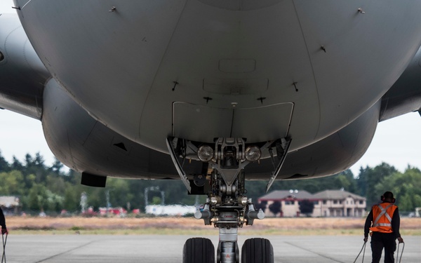 A ground-level view beneath a large commercial or military aircraft on a tarmac, showing the nose landing gear in sharp focus at center frame. Two ground crew members wearing high-visibility orange safety vests are visible on either side, handling yellow wheel chocks attached to ropes. The image is taken from a low angle looking forward along the aircraft's centerline, with trees and airport buildings visible in the background.
