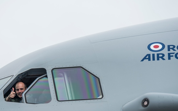 A Royal Air Force aircraft is shown in close-up, with a pilot or crew member leaning out of an open cockpit side window giving a thumbs-up gesture. The aircraft fuselage is painted in light grey and bears the official RAF roundel logo alongside the 'Royal Air Force' text in blue. The image appears to be taken on an overcast day, likely on an airfield.