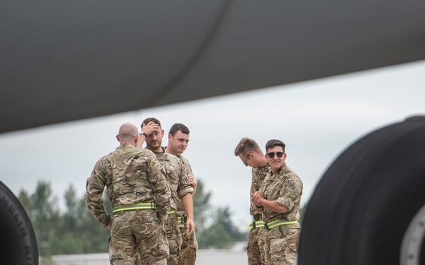 A group of five military personnel in camouflage uniforms are gathered on an aircraft tarmac, photographed from beneath a large aircraft between two sets of landing gear. The personnel appear to be ground crew or aircrew engaged in a briefing or casual conversation. Yellow wheel chocks are visible securing the aircraft's landing gear.