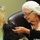 An elderly woman wearing a white sequined hat and black-framed glasses is gently petting a small black dog. A younger woman with curly blonde hair is present on the left side, appearing to hold or assist with the dog. The setting appears to be an indoor facility, possibly a care or assisted living environment.