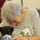 An elderly woman with white/gray hair is leaning forward to interact closely with a small black and white puppy or kitten held in her lap. She is wearing a black and white striped shirt with a decorative patch featuring a tree and giraffe motif. A younger person in a green shirt is partially visible on the left side of the frame.