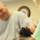 A middle-aged bald man wearing a gray t-shirt is smiling warmly while petting a small dark-colored dog, likely a Yorkshire Terrier or similar breed. A second person with blonde curly hair wearing a green shirt is partially visible on the right side, also interacting with the dog. The setting appears to be an indoor room with a round wall clock visible in the background.