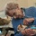 An elderly woman wearing a blue cable-knit cardigan and supplemental oxygen nasal cannula is seated in a wheelchair, holding and looking down affectionately at a small Chihuahua. The woman wears gold-framed glasses, a pearl necklace, and a ring, and appears to be in an indoor care facility setting. The interaction suggests a pet therapy or visitation session.