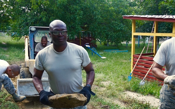 Several military personnel in U.S. Air Force camouflage uniforms are performing manual labor at what appears to be a community service or humanitarian assistance project. They are clearing debris, moving rocks and broken concrete, and operating a small skid-steer loader in an outdoor area near a colorful playground structure. The work appears to be site preparation or cleanup at a facility, possibly a school or community center.