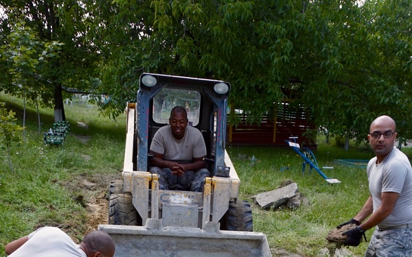 Several individuals in military-style gray t-shirts and camouflage pants are engaged in construction or demolition work outdoors. A skid-steer loader (bobcat) is positioned in the center while workers manually remove broken concrete and debris from a trench or pathway. The work appears to be taking place in a park or recreational area with trees and grass visible in the background.