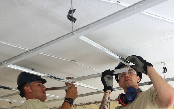 Two military personnel in US Air Force ABU (Airman Battle Uniform) camouflage are installing a suspended ceiling grid system inside what appears to be a children's room or facility. They are standing on aluminum step ladders and working with metal ceiling track rails. A third person in civilian or partial uniform is visible in the background.
