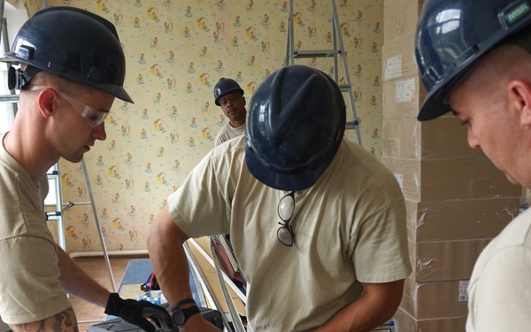 Three military personnel wearing hard hats and tan/camouflage uniforms are engaged in construction or renovation work inside a building with floral wallpaper. They are working at a table covered with tools and equipment, appearing to cut or assemble metal components. A fourth person is visible in the background near a ladder.