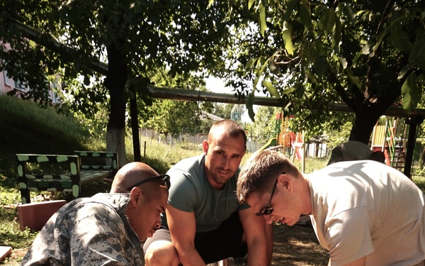 Three men are working together outdoors in what appears to be a community service or construction project. Two individuals are wearing military camouflage uniforms (US Army ACU pattern and what appears to be Air Force ABU pattern), while a third man in civilian clothing assists them. They are working in sandy/dirt ground, apparently setting or positioning a wooden post or board near what looks like a playground area.