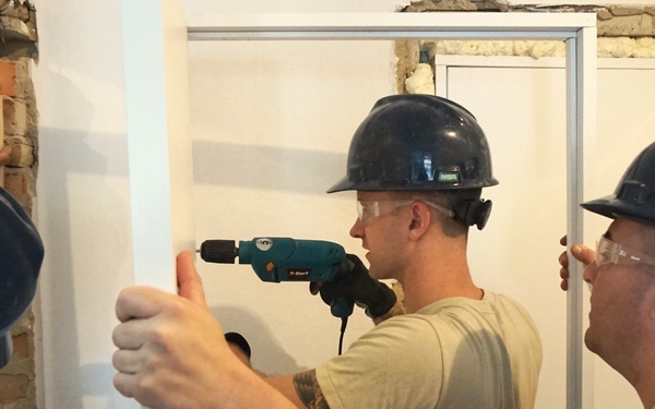 Three workers wearing blue hard hats and safety glasses are installing a door frame in a building undergoing renovation. The middle worker is using a power drill to secure the white door frame, while the other two workers hold it in position. The surrounding wall shows exposed brick, indicating active construction or remodeling work.