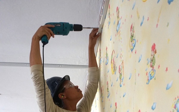 A person wearing military-style camouflage pants and a tan long-sleeve shirt is using a corded power drill at the ceiling-wall junction of a room. The walls are covered with children's wallpaper featuring clown and moon motifs on a yellow background. The individual is standing on a step or platform and appears to be performing renovation or installation work.