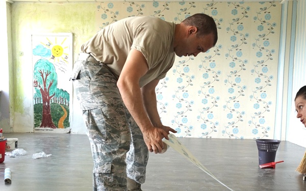 A military servicemember in camouflage uniform works alongside two civilian women on a renovation or installation project inside what appears to be a children's classroom or daycare facility. The group is measuring and cutting what looks like wallpaper or flooring material laid out on the floor. The setting suggests a community service or humanitarian assistance project.
