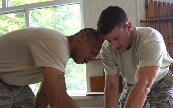 Two individuals wearing US Air Force Airman Battle Uniforms (ABUs) are collaborating on a construction or renovation task indoors. They are measuring or marking a ceiling tile on a table, with one person pointing at a specific location on the tile. The setting appears to be a building undergoing repair or improvement work.