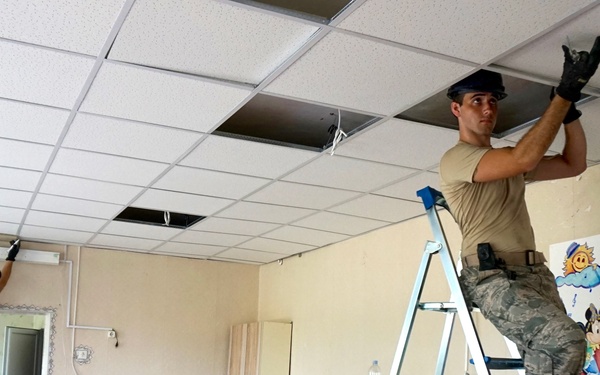Two military personnel in U.S. Air Force camouflage uniforms are working on a drop ceiling renovation inside what appears to be a children's facility or classroom. They are standing on aluminum ladders, removing or installing ceiling tiles while the room's furniture — small blue and wood chairs — is stacked and piled in disarray below. A colorful children's mural featuring Mickey Mouse and Minnie Mouse is visible on the wall, suggesting this is a school or daycare renovation project.