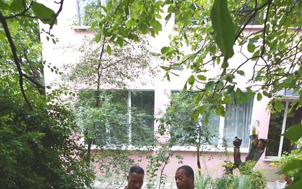A group of individuals in military camouflage uniforms observe a civilian worker laying decorative tiles in a courtyard. The work appears to be a construction or renovation project, likely a humanitarian or community assistance mission. A colorful traffic light prop or decoration is visible nearby, and a pink building is seen in the background.