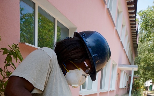 A person wearing U.S. Air Force Airman Battle Uniform (ABU) camouflage pants, a gray t-shirt, black hard hat, safety glasses, N95 respirator mask, and black gloves is operating a yellow miter saw to cut what appears to be laminate or vinyl flooring planks. The work is being performed outdoors adjacent to a pink stucco building, suggesting a construction or renovation project. Additional power tools, including a blue Bort jigsaw, are visible on the work surface nearby.
