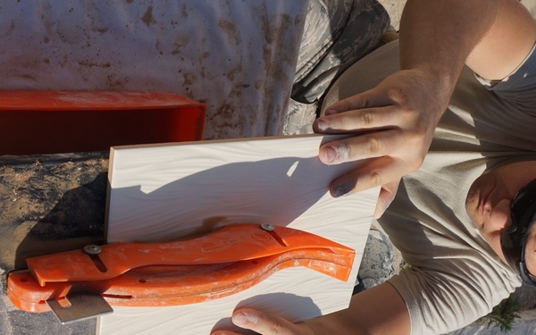 A young man is working outdoors using a manual tile cutter with an orange handle to score or cut a ceramic or porcelain tile. He is leaning over a work surface covered with a plastic sheet, carefully positioning the tile on the cutting tool. The setting appears to be an outdoor renovation or tiling project.