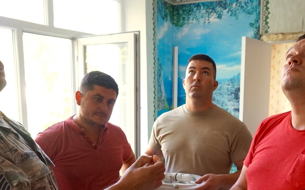 Four men are gathered inside what appears to be a building under renovation or repair. Two individuals are wearing military uniforms (one in US Air Force camouflage with a Master Sergeant chevron patch, one in tan/OCP-style uniform), while two are in civilian clothing. They are examining ceiling light fixtures, with at least two men looking upward, apparently planning or discussing installation.