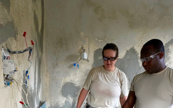 Three individuals in U.S. Air Force digital camouflage uniforms are engaged in construction or renovation work inside a partially finished room. One person is crouching and installing light blue wall tiles while two others stand and observe. The setting appears to be a humanitarian assistance or civil engineering project.