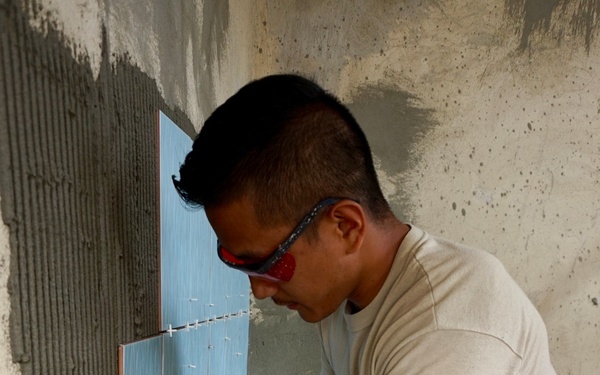 A person wearing US Air Force ABU (Airman Battle Uniform) camouflage pants and a tan t-shirt is installing light blue ceramic wall tiles in what appears to be a construction or renovation project. The individual is crouched down, pressing tiles against a mortar-covered wall with both hands, using tile spacers to maintain uniform grout lines. The setting appears to be an unfinished interior space with bare concrete floors and walls.