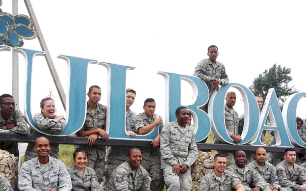 A large group of U.S. Air Force personnel in ABU (Airman Battle Uniform) camouflage are posing for a group photo in front of a large decorative sign reading 'HULBOACA'. The group includes both men and women of diverse backgrounds, arranged in multiple rows with some sitting, some standing, and one individual perched on top of the sign letters. The location appears to be Hulboaca, Moldova, likely during a military exercise or community engagement mission.
