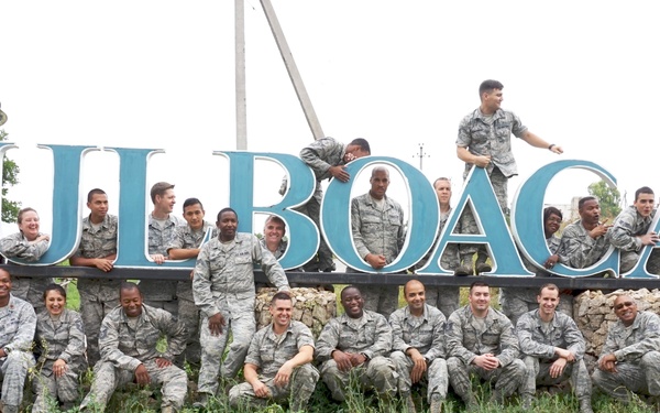 A large group of U.S. military personnel in Air Force Airman Battle Uniform (ABU) camouflage are posing for a group photo in front of a large town entrance sign reading 'HULBOACA'. The sign features a decorative grape cluster motif, suggesting this is a village in Moldova known for viticulture. Some personnel are seated on the ground, standing behind the sign, and two individuals are climbing on top of the letters.