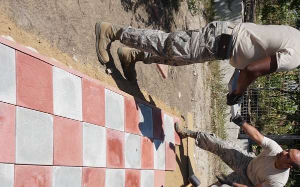 Three individuals in military-style uniforms are laying a checkerboard-patterned tile floor consisting of alternating red and white/gray square tiles. The work appears to be an outdoor construction or improvement project, with the tiles being set into a prepared ground surface. One person is using a rubber mallet to set the tiles while the others assist with placement and alignment.
