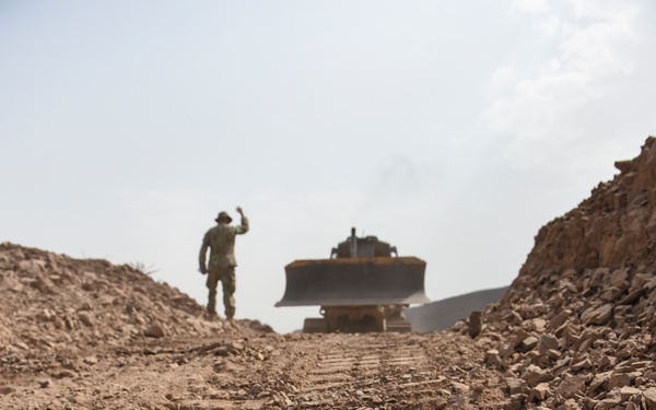 A ground-level perspective of a rocky earthworks site showing a bulldozer actively grading or cutting through arid terrain. A person in military-style camouflage uniform and hat stands on an elevated mound to the left, raising one arm as if signaling to the bulldozer operator. The foreground is dominated by churned rocky soil with visible track impressions from heavy machinery.