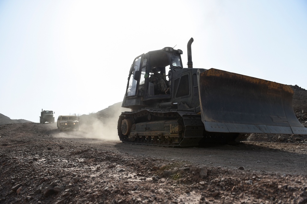 A military bulldozer with a large front blade is operating on a dusty, rocky terrain, likely in an arid or desert environment. Behind it, two military trucks follow along the same unpaved road, kicking up dust. The scene suggests a military engineering or construction operation in a remote location.