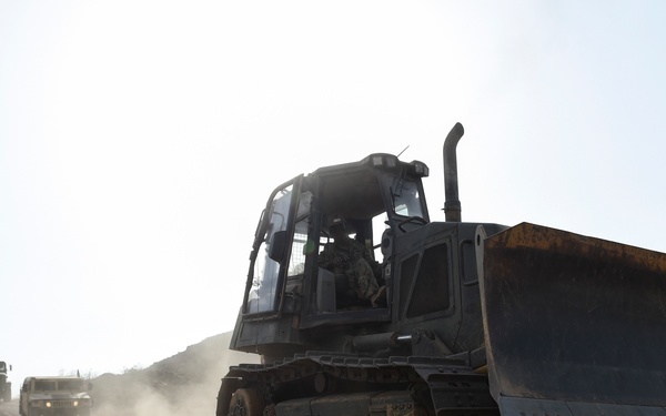 A military bulldozer with a large front blade is operating on a dusty, rocky terrain, likely in an arid or desert environment. Behind it, two military trucks follow along the same unpaved road, kicking up dust. The scene suggests a military engineering or construction operation in a remote location.