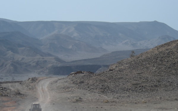 The image shows a dusty unpaved road cutting through an arid, rocky desert landscape with large spoil heaps or tailings piles visible on either side, suggesting an active mining or quarrying operation. Several vehicles, including what appears to be a flatbed truck carrying equipment and a smaller vehicle, are traveling along the dirt road raising dust clouds. Barren mountain ranges are visible in the background under a hazy sky.