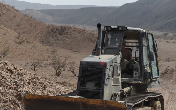 A military bulldozer painted in olive drab/gray camouflage is being operated in a rocky desert terrain. The tracked vehicle, equipped with a front blade, is actively grading or clearing a rocky path through an arid, mountainous landscape. A single operator is visible inside the enclosed cab.