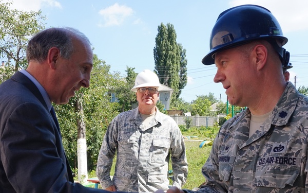A civilian in a dark suit is shaking hands with a U.S. Air Force servicemember wearing ABU (Airman Battle Uniform) and a dark hard hat at what appears to be a construction or project site. Several other U.S. Air Force personnel in ABUs are visible in the background, some also wearing hard hats, suggesting an official site visit or inspection. The setting appears to be an outdoor location with residential buildings and trees visible in the background.
