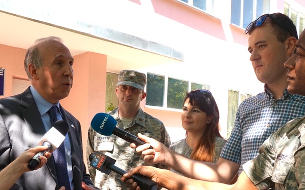 A press briefing or media interview is taking place outdoors in front of a pink building, likely in Eastern Europe based on visible signage in Romanian. A man in a dark suit is being interviewed by reporters holding microphones, while U.S. Air Force personnel and civilians stand nearby. The setting appears to be an official visit or humanitarian/military cooperation event.