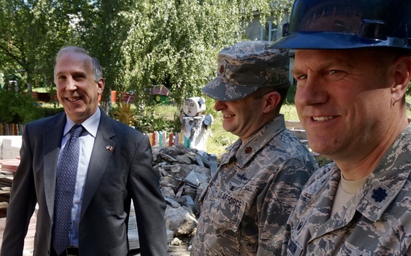 Three individuals stand at what appears to be a construction or renovation site outdoors. Two are wearing US Air Force ABU (Airman Battle Uniform) camouflage uniforms, with one also wearing a blue hard hat. The third person is a civilian in a dark suit with a tie, wearing a small lapel pin.