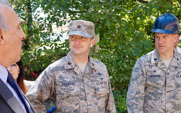 A civilian in a suit is meeting with several U.S. Air Force personnel in ABU (Airman Battle Uniform) camouflage. Three of the military members are wearing blue hard hats, suggesting an active construction or engineering project site. Name tapes visible on the uniforms read 'PAQUETTE,' 'SWANSON,' and 'BULLER.'