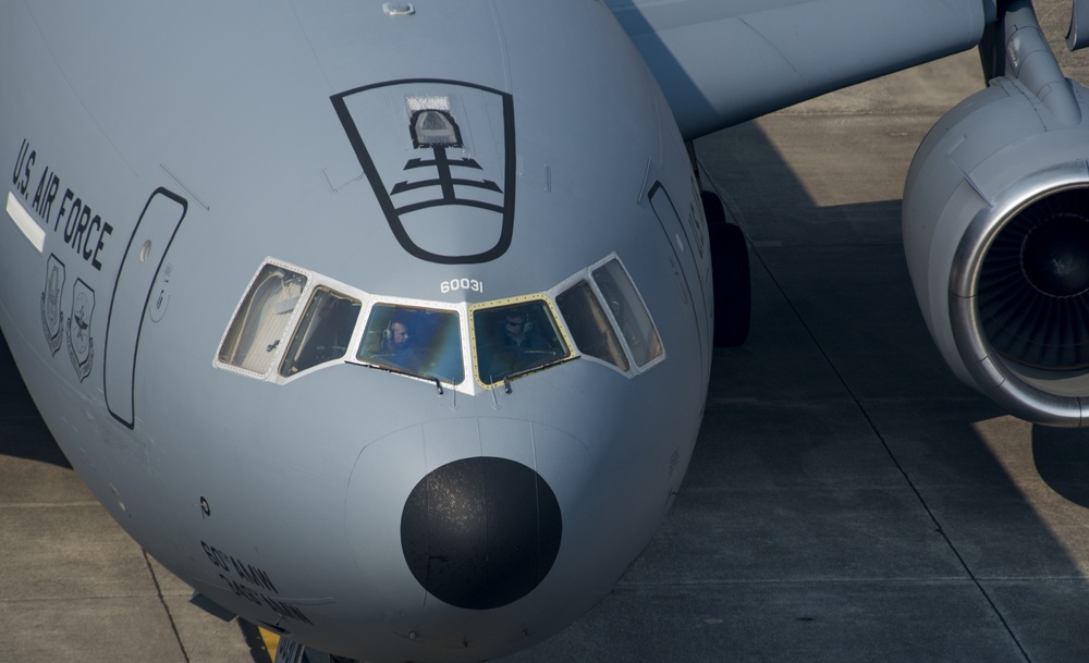 A close-up frontal view of a U.S. Air Force C-17 Globemaster III military transport aircraft on a tarmac. The aircraft's nose section is prominently featured, showing the cockpit windows with crew members visible inside. The serial number 60031 is visible below the cockpit, and a unit insignia shield is displayed on the fuselage.