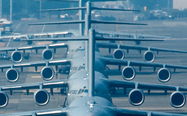 Multiple U.S. Air Force C-17 Globemaster III cargo aircraft are lined up on a taxiway or flight line, creating a dramatic compressed perspective due to telephoto lens compression. The aircraft are painted in standard military gray livery with 'AIR FORCE' and 'US AIR FORCE' markings clearly visible. A single ground crew member in a red/orange high-visibility vest walks in front of the lead aircraft.