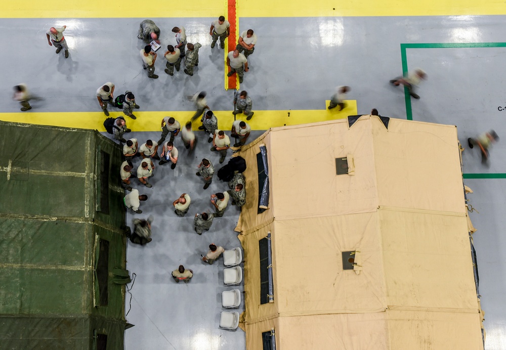 An aerial/overhead view of what appears to be a military indoor facility, likely a large hangar or gymnasium, with numerous personnel in military uniforms gathered between two large military tents or shelters. The floor features painted yellow and green boundary lines, suggesting a structured operational or training environment. Some figures appear motion-blurred, indicating movement during a longer exposure photograph.
