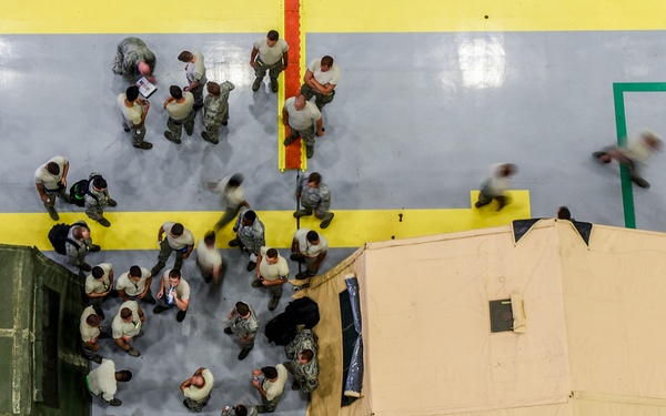An aerial/overhead view of what appears to be a military indoor facility, likely a large hangar or gymnasium, with numerous personnel in military uniforms gathered between two large military tents or shelters. The floor features painted yellow and green boundary lines, suggesting a structured operational or training environment. Some figures appear motion-blurred, indicating movement during a longer exposure photograph.