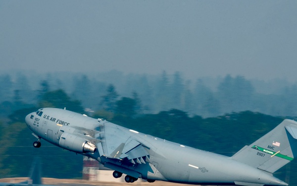 A U.S. Air Force C-17 Globemaster III military transport aircraft is captured during takeoff rotation from a runway. The aircraft bears tail number 21107 and AMC (Air Mobility Command) markings with a green stripe on the tail. A second military aircraft, appearing to be a C-130, is visible on the tarmac in the background.
