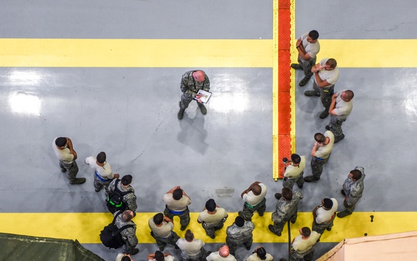 An aerial/overhead view of a military briefing or formation taking place inside what appears to be a large hangar or indoor facility. A group of approximately 25-30 uniformed military personnel are gathered in a loose formation facing a single individual who appears to be addressing or briefing them. Two military field tents or shelters (one olive drab, one tan/desert) are visible at the bottom corners of the frame.