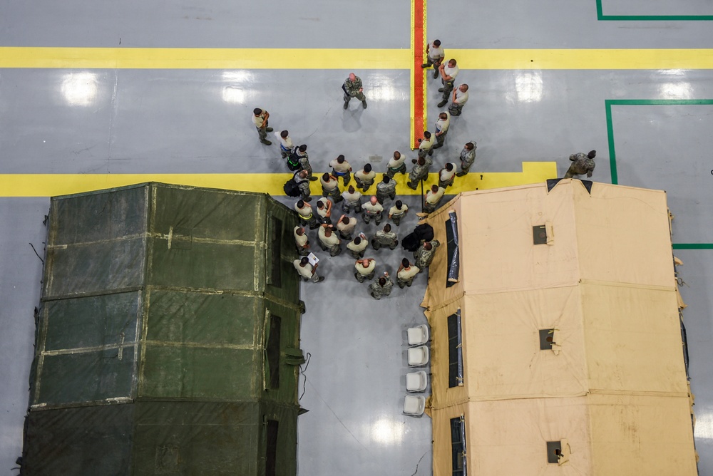 An aerial/overhead view of a large indoor facility, likely a military hangar or gymnasium, showing a group of uniformed personnel gathered in a briefing or formation between two military field tents. One tent is olive green and the other is tan/desert colored, both set up on a polished gray floor marked with yellow and green lines. A single individual appears to be addressing the assembled group.