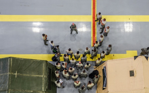 An aerial/overhead view of a large indoor facility, likely a military hangar or gymnasium, showing a group of uniformed personnel gathered in a briefing or formation between two military field tents. One tent is olive green and the other is tan/desert colored, both set up on a polished gray floor marked with yellow and green lines. A single individual appears to be addressing the assembled group.