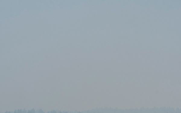 A military airfield ramp is densely packed with multiple large military transport aircraft, consistent with C-17 Globemaster III airlifters, parked in close proximity. The atmosphere is heavily obscured by haze or smoke, significantly reducing visibility and giving the scene a muted, gray-blue tone. The setting appears to be a large military air base with tree lines and infrastructure visible in the background.