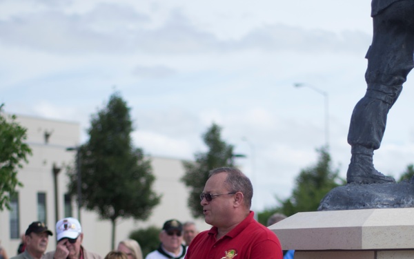 A man in a red polo shirt with sunglasses is speaking to a gathered crowd at what appears to be a military memorial dedication or ceremony. He is standing beside a stone monument bearing a bronze plaque inscribed 'In Memory of our Fallen Comrades' with names listed, topped by a bronze soldier statue. The audience includes men and women, some wearing name badges and caps, gathered outdoors on a cloudy day.