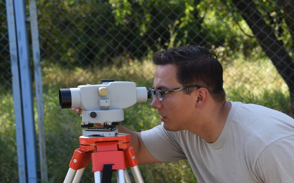 A young man wearing glasses and a beige t-shirt is looking through a surveying level instrument mounted on a white and orange tripod. He is holding a notepad and what appears to be a yellow folder in his other hand. The setting is an outdoor area with grass, trees, and a chain-link fence visible in the background.