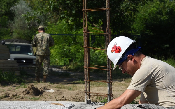 A military service member wearing camouflage uniform and a white hard hat is crouching at a construction site, using a tape measure along a concrete slab edge. A second uniformed individual stands in the background near a parked vehicle. The scene appears to be a military engineering or construction project in a wooded area.