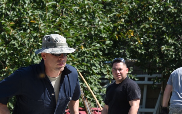 The image shows a group of individuals, some wearing military-style camouflage uniforms, engaged in a construction or community service project outdoors. The foreground figure is carrying two concrete cinder blocks while wearing work gloves and a camouflage boonie hat. The setting appears to be a work site near a building surrounded by dense green foliage.