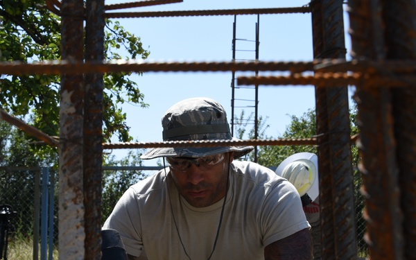 A worker in military-style clothing and a bucket hat is photographed through a rebar grid structure at a construction site. He appears to be working at ground level, handling construction materials. A second worker wearing a white hard hat is visible in the background.